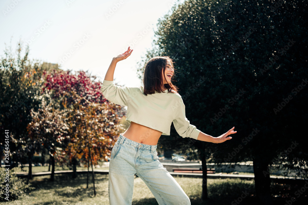 Naklejka premium Happy young woman dancing in a sunlit park wearing a casual crop top and jeans, carefree summer lifestyle portrait expressing joy and movement