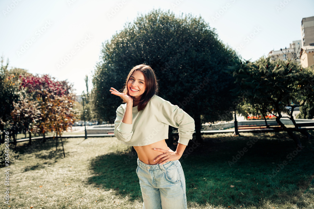 Fototapeta premium Smiling young woman in cropped sweatshirt and jeans posing playfully in a sunny city park