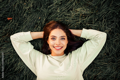 Smiling young woman lying on grass  relaxed outdoors with hands behind head enjoying sunny day and natural green surroundings