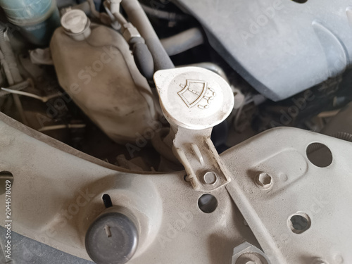 Close up of a cars dusty engine bay, focusing on the white plastic windshield washer reservoir cap. The cap features a standard windshield wiper symbol surrounded by dirty engine components.
