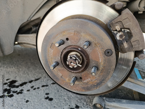 Close-up shot shows a car's disc brake assembly during maintenance. The wheel is removed, exposing a rusty wheel hub with studs, a shiny brake rotor, a caliper, and a jack lifting the vehicle