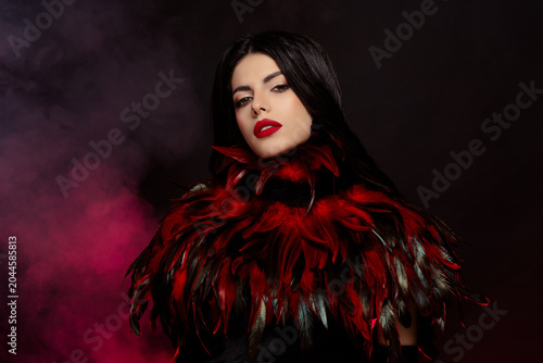 Elegant woman in a feathered red costume with striking makeup surrounded by dramatic smoky lighting.