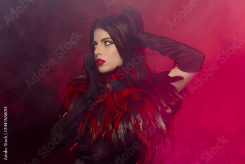 Elegant young woman in a luxurious feather costume posing dramatically against a smoky background