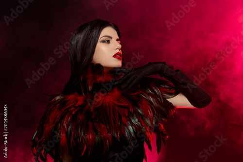 Elegant young woman with a feathered costume and bold makeup in a smoky dramatic red backdrop