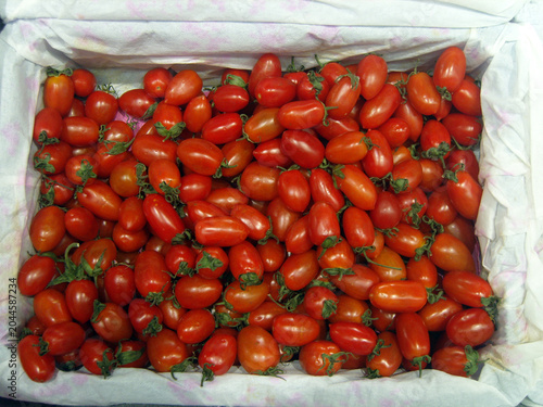 Top-down view of white paper-lined crate filled with fresh, vibrant red cherry tomatoes. The ripe, oblong tomatoes still have their green stems attached, creating a colorful and textured harvest scene