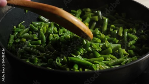 Hand with wooden spoon stirring green bean pods in frying pan