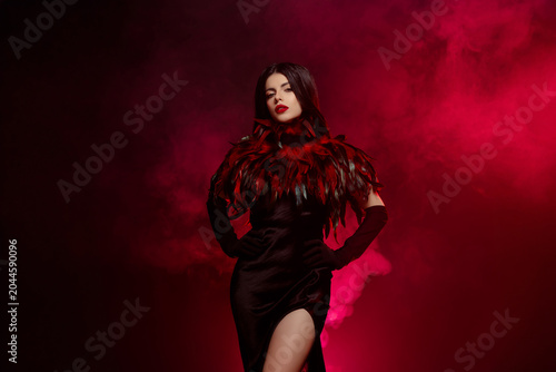 Elegant brunette woman in a stylish feathered gown posing in a vibrant, smoky setting for a glamorous fashion shoot