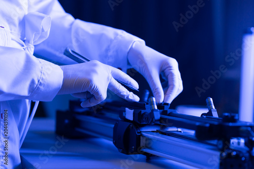 Person in lab coat and gloves operates tools on equipment in dark room