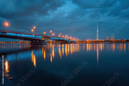 Vibrant Cityscape at Dusk Featuring a Bridge Illuminated with Blue and Orange Lights Reflected in Calm Water Under a Cloudy Sky