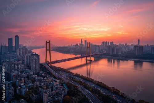 Vibrant Cityscape at Dusk With a Suspended Bridge Over a Wide River Illuminated by City Lights and a Fiery Sunset Sky Showing Apartment Buildings and Skyscrapers