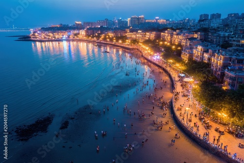 Vibrant Coastal Cityscape at Dusk with Sparkling Lights Reflecting on Calm Ocean Waters Crowds of People Stroll Along the Beachfront Promenade Enjoying the Evening Ambiance