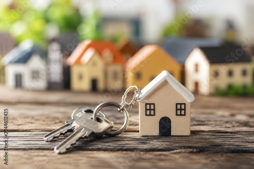 House model with keys on wooden table and blurred miniature houses in background.
