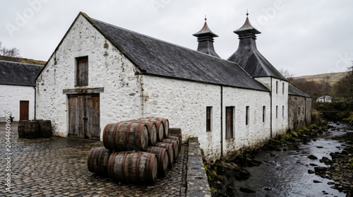 Historic distillery buildings with aging whisky barrels stacked outside by a flowing stream