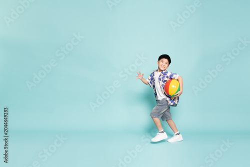 Full body of a happy Asian boy running while holding a colorful beach ball. Cheerful young kid in a summer shirt playing and smiling, isolated on a green studio background with large copy space