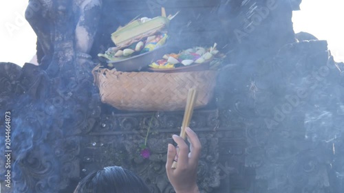 Balinese Hindu offering ceremony, with a woman placing flowers and incense on a ceremonial tray. Rising smoke creates a spiritual atmosphere during a temple ritual in Bali, Indonesia