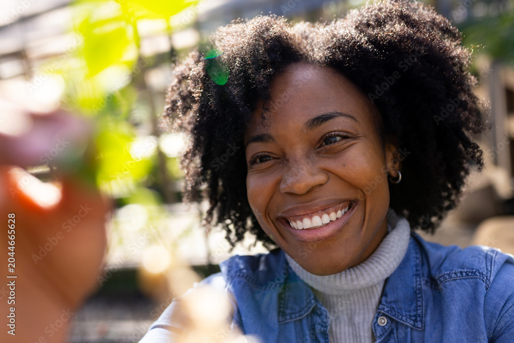 Obraz premium Mid-adult African American woman smiling reaching for green leaves in sunny garden in denim jacket