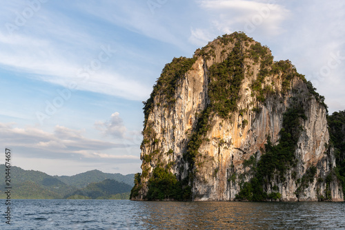 Cuadro en lienzo Majestic limestone cliff face, lush green jungle, and calm water under a soft blue sky with wispy clouds