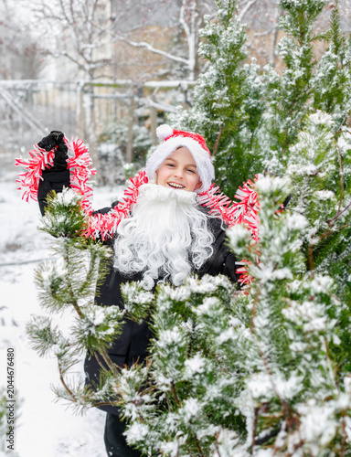 Child dressed as Santa with white beard red tinsel garland standing outdoors in snowy winter garden