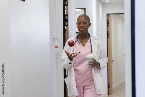 Obraz na płótnie African woman in pink scrubs walking clinic hall tossing red apple holding coffe