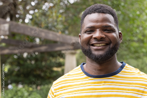 African American man standing, smiling, wearing striped yellow tee with navy collar by pergola