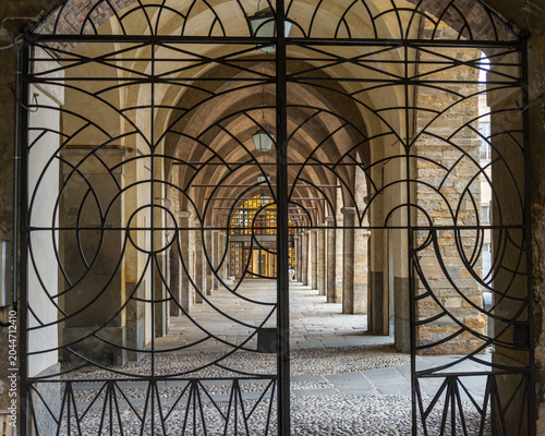 Gate made of wrought iron in the historical centre of the city of Bergamo, Lombardy, Italy. In the backgrouond the medieval arcades of a courtyard, with cobblestone ground.