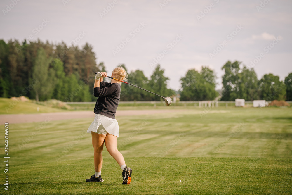Obraz premium Female golfer in black sweater and white skirt swings golf club on green course with trees and open field in the background under a clear sky