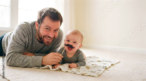 Smiling father playing with baby using funny dad jokes mustache prop
