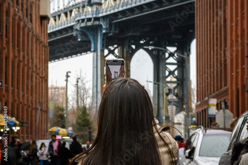 Female tourist taking photo of Manhattan Bridge with her phone in DUMBO, Brooklyn, New York.