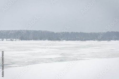 Schilderij op canvas Quiet winter landscape featuring a frozen rive rwith snow-covered surface, distant snowy forest and a soft foggy horizon