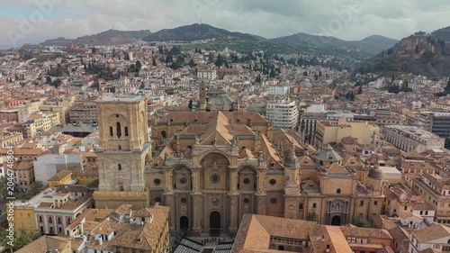 Huge Renaissance Cathedral and Moorish Alhambra complex stand in Granada, Spain. Terracotta roofs fill dense urban cityscape of ancient Andalusian town. 4K aerial of distant Spanish mountains