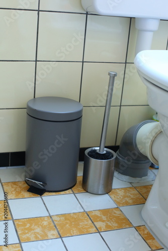 Modern bathroom trash can and toilet brush standing next to a toilet in a tiled bathroom. Household hygiene, sanitation and bathroom cleaning concept