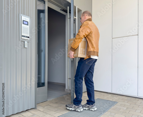 Man holds cardboard box at a modern apartment entrance. He stands by an open glass door and access panel in daylight. Delivery and relocation service concept..