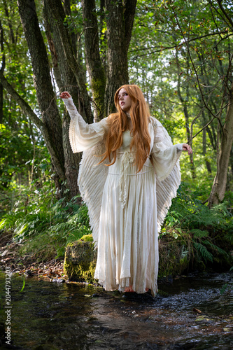Young red-haired woman with angel wings and a long white dress stands by a stream, playing with her hands