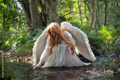 Young red-haired woman with angel wings and a long white dress, sitting by a stream and playing with the water
