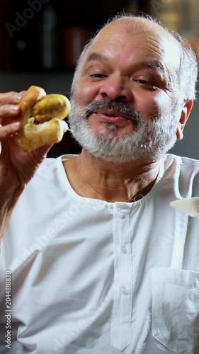 Senior Indian man eating vada pav with tea at home table, Asian foodie old man expressing immense joy and satisfaction while enjoying spicy Mumbai street food snack in modern Indian kitchen setup