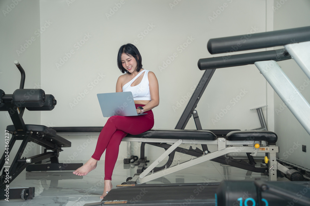Fototapeta premium Smiling young Asian woman in sportswear sitting on a fitness bench and using a laptop, working remotely, checking her workout plan, or managing an online fitness business in a gym