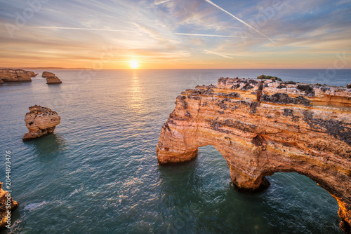 Sunrise view of Praia da Marinha beach in Algarve, Portugal, with a limestone sea arch, Atlantic water, warm sky, and rugged coastal cliffs.