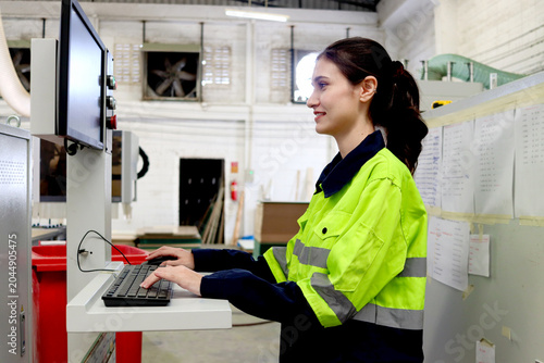 Beautiful female mechanic engineer in safety protective clothes uses a computer to operate machinery in a manufacturing plant. Industrial woman worker working with machine in industry factory.