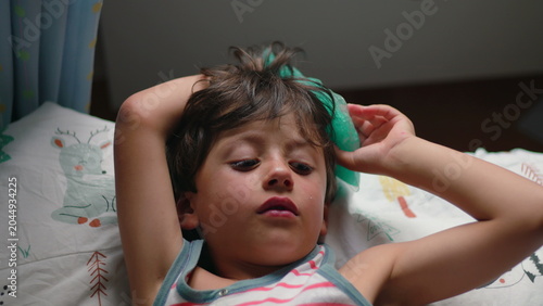 Bild auf Leinwand Child lying on bed holding ice pack against forehead with tired eyes expressing