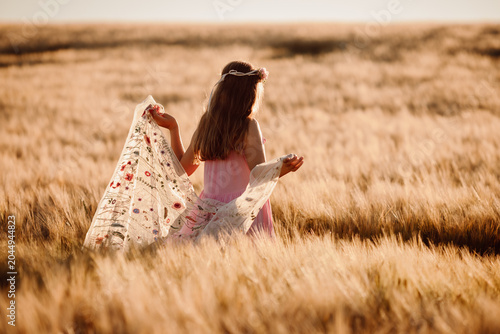 Quadro su tela Girl walking through golden wheat field trailing floral shawl behind her