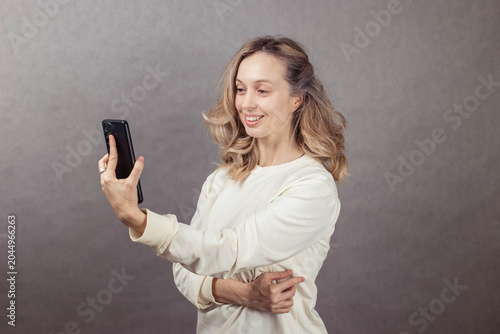 Smiling woman with wavy hair is taking a selfie with a smartphone, showcasing a joyful expression against a neutral textured background, capturing a moment of happiness and connection