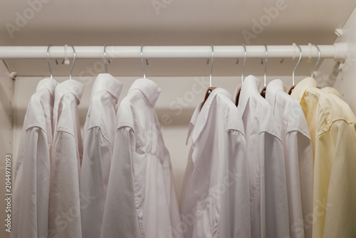 row of clean white and yellow button down shirts hanging on a rack inside a closet in bedroom at apartment. Minimalist wardrobe background representing individuality, organization, and standing out.