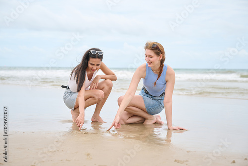 Obraz na plátně Young women are squatting on a wet sandy beach, looking down at something they a