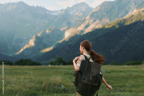 Fotografija A young woman with a backpack walks through a sunlit grassy field toward distant
