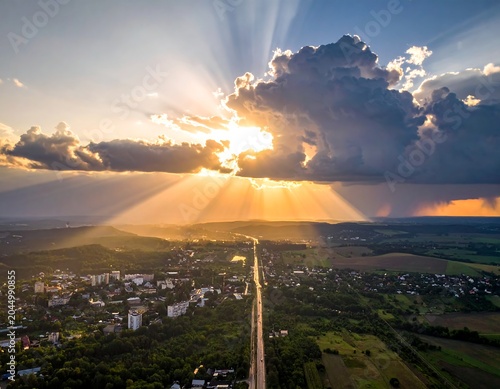 Aerial view of a serene landscape with a road cutting through a small town surrounded by lush green fields and trees under a cloudy sunset sky