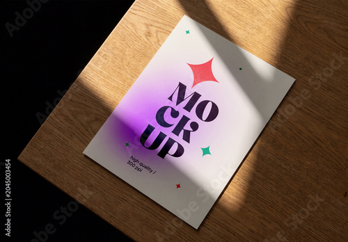 High-angle shot of a blank flyer or poster mockup lying on a wooden table with harsh light and shadows cast across the surface