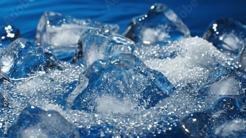 Macro close-up of crystal clear ice chunks and crushed granular ice resting on a blue surface with visible water droplets and reflections under bright studio lighting.