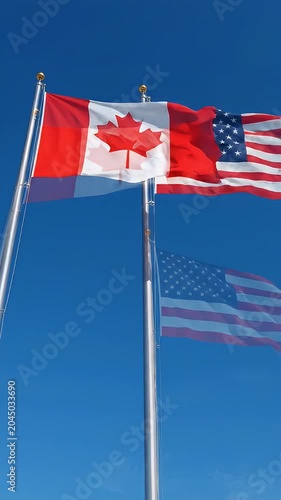 Canadian and American Flags Waving Together in Blue Sky - The Canadian flag and the American flag are shown flying side-by-side on flagpoles against a clear blue sky.