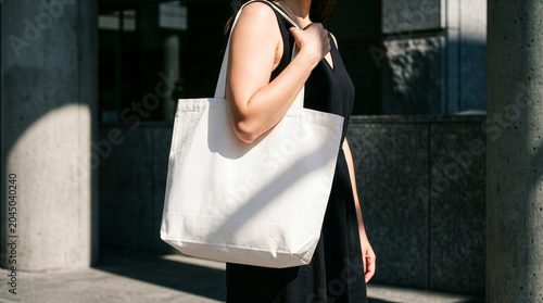 Fotografie A woman wearing a black dress carries a blank white canvas tote bag in a sunny urban environment with hard shadows