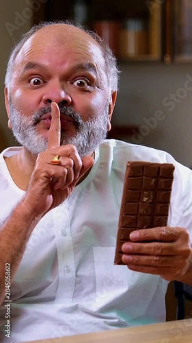 Senior Indian man secretly eating chocolate brick in modern kitchen, Asian elderly foodie sneaking thick chocolate slab with mischievous expression, enjoying sweet treat alone at home